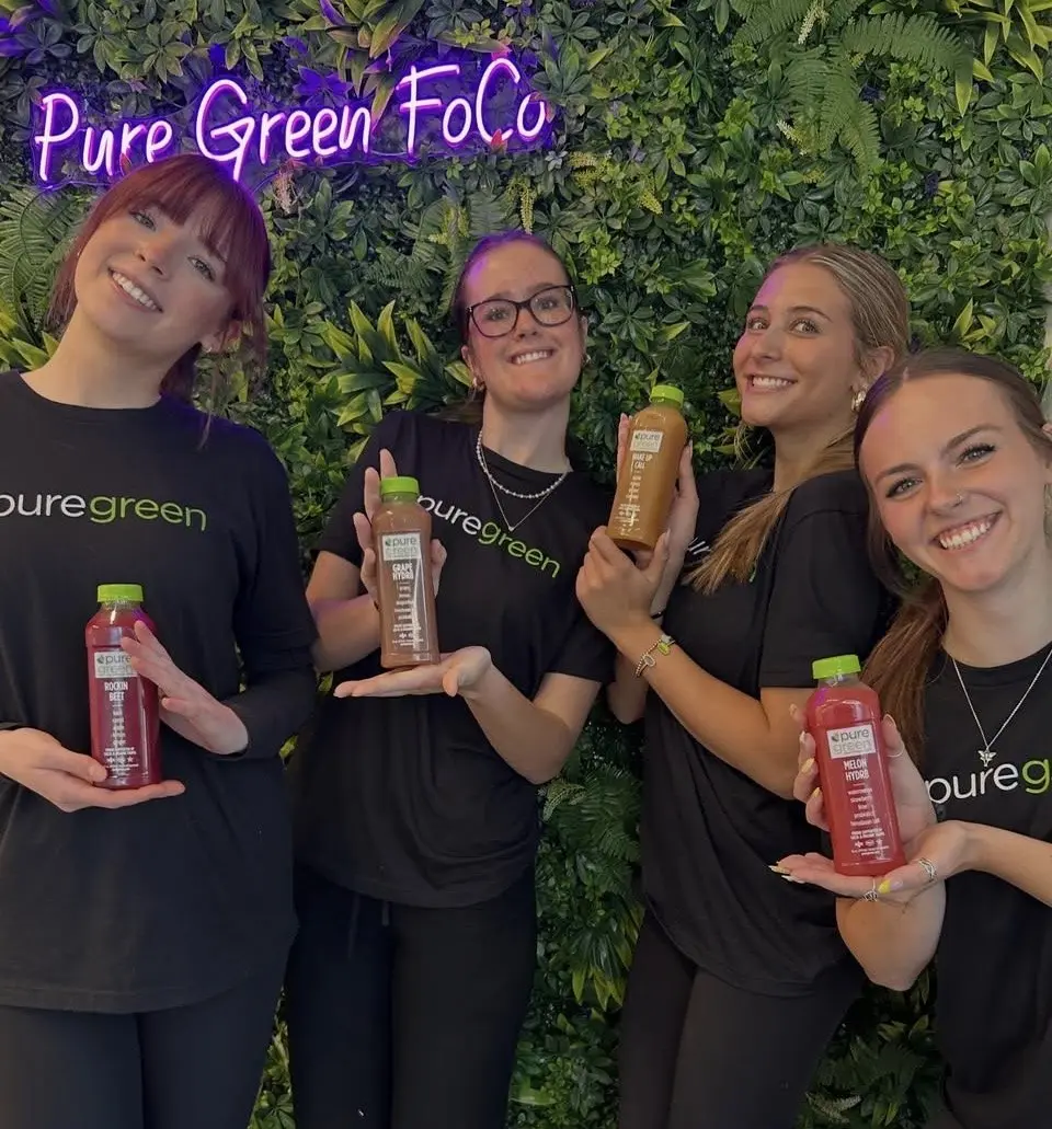 The Pure Green Fort Collins team holding cold-pressed juices by the greenery wall